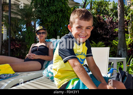 Young boy with with huge smile poolside at a holiday resort. His good looking mother in her forties looks on relaxing. Stock Photo