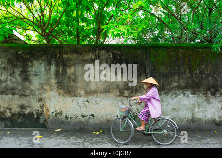 Vietnam woman cycling, view of a senior Vietnamese woman wearing a conical hat and dressed in pink cycling along a street in Hoi An, Central Vietnam. Stock Photo