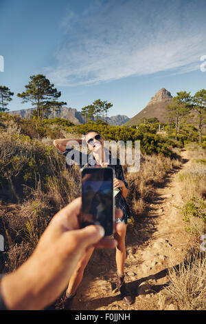Young woman posing to mobile phone camera. Man hand holding a smart phone taking pictures on a young woman. Couple hiking in cou