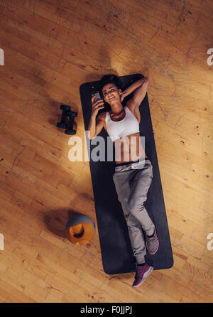 Top view of woman relaxing on yoga mat using mobile phone. Fitness female taking a break lying on exercise mat at gym reading te Stock Photo