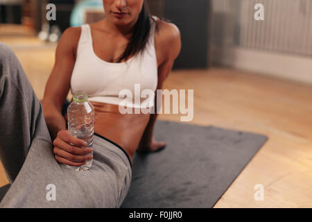 Cropped shot of relaxed young woman holding water bottle. Fitness woman in sports wear sitting on exercise mat at gym. Stock Photo