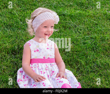 Little beautiful girl sitting on the green grass outdoors in summer garden. Stock Photo