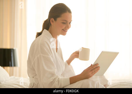 Woman in bathrobe drinking coffee and using digital tablet in bedroom Stock Photo
