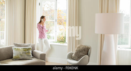 Woman drinking coffee and talking on cell phone at living room window Stock Photo