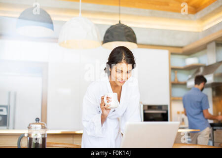 Woman in bathrobe drinking coffee and using laptop in kitchen Stock Photo