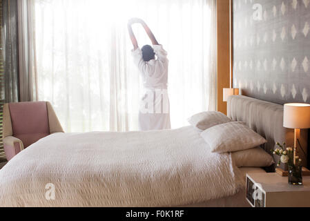 Woman in bathrobe stretching with arms raised in bedroom Stock Photo