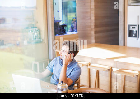 Man talking on cell phone at laptop in kitchen Stock Photo