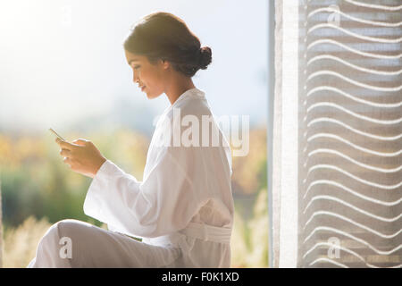 Woman in bathrobe texting with cell phone in sunny doorway Stock Photo