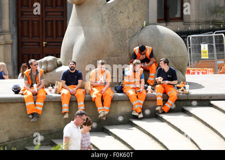 A group of workmen, construction workers, sitting side by side, wearing bright overalls, take their lunch, sat on a shaded wall in a busy city centre Stock Photo