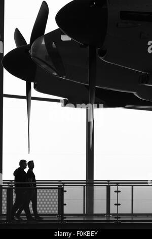 Two visitors at the imperial war museum, Duxford, walk past a de Havilland Mosquito that hangs from the roof of AirSpace. Stock Photo