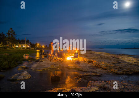 Family and friends warm up by a bonfire under a moon-lit night as high tide slowly trickles inland. Stock Photo