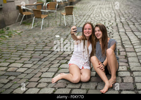 Two hilarious teenage girls are doing selfie on the phone sitting on the pavement. Stock Photo