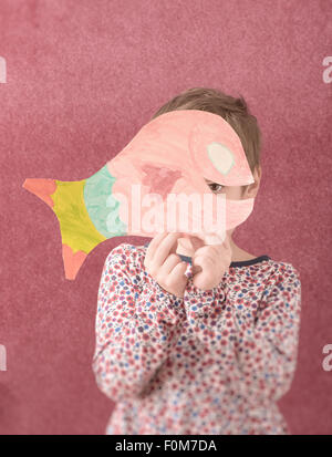 Portrait of little girl in dress holding a cut out drawing of a fish in front of her face. She is shy and hiding behind it. Stock Photo