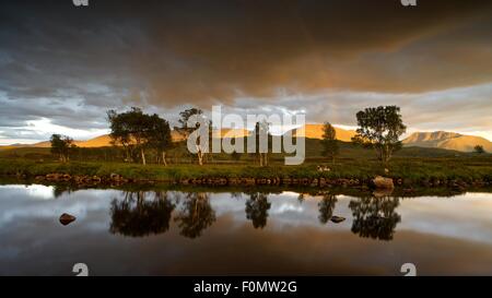 Golden light hits the hills above Rannoch moor from the setting sun with a reflection of dark clouds and a rainbow in Loch Ba Stock Photo