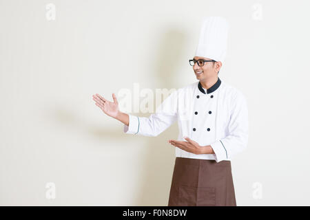 Portrait of handsome Indian male chef in uniform hands showing something and smiling, standing on plain background with shadow, Stock Photo