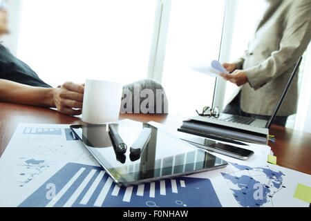 business documents on office table with smart phone and digital tablet and stylus and two colleagues discussing data in the background Stock Photo
