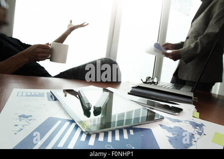 business documents on office table with smart phone and digital tablet and stylus and two colleagues discussing data in the background Stock Photo