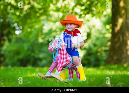 Little boy dressed up as cowboy playing with his toy rocking horse in a summer park. Kids play outdoors. Stock Photo