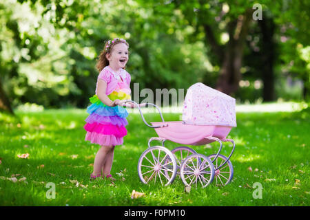 Little girl pushing toy stroller with bear. Toddler kid in pink dress playing with doll buggy. Kids birthday party. Stock Photo