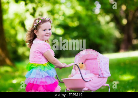 Little girl pushing toy stroller with bear. Toddler kid in pink dress playing with doll buggy. Kids birthday party. Stock Photo