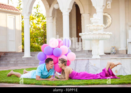 Happy young couple lying down on the lawn in front of their villa and holding colorful balloons Stock Photo