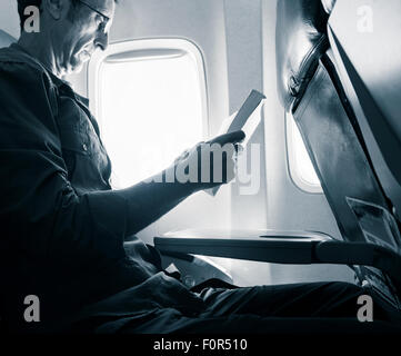 Mature man sitting in window seat on airplane reading a book. Stock Photo