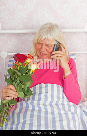 Happy woman in pink pajama sitting in bed with a bunch of roses and talking on the phone Stock Photo