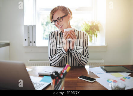 Happy successful businesswoman taking a break to drink a mug of coffee as she reads information on her laptop screen with a smil Stock Photo