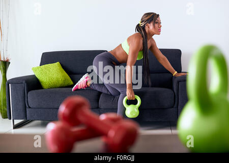Young adult african american woman in sports clothing at home, doing domestic fitness and training shoulders with weight on sofa Stock Photo