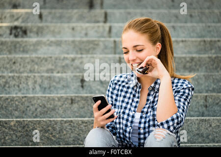Teenager - young woman eating chocolate in street and looking in phone Stock Photo