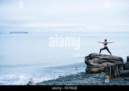Mature woman practicing yoga pose whilst standing on large driftwood tree stump on beach Stock Photo