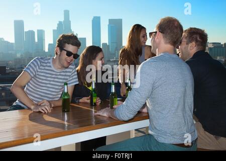 Six adult friends talking and drinking beer at rooftop bar with Los Angeles skyline, USA Stock Photo
