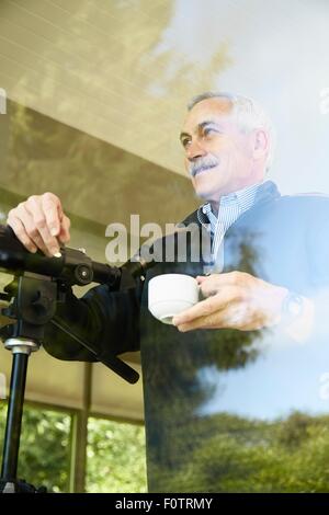Senior man at home, drinking coffee, using telescope through window Stock Photo