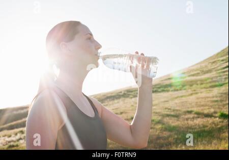 Young female runner drinking bottled water on sunlit hillside