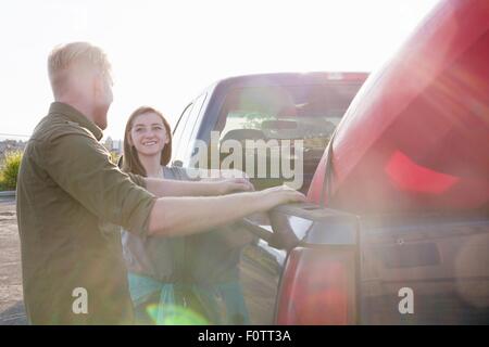 Young couple leaning against pick up truck, looking at each other smiling, lens flare Stock Photo