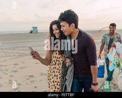 Group of friends walking along beach, young couple looking at smartphone