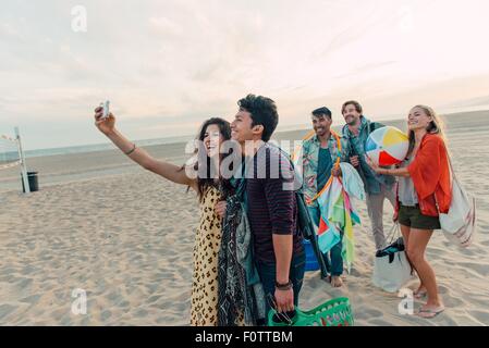 Group of friends walking along beach, young couple taking self portrait with smartphone Stock Photo