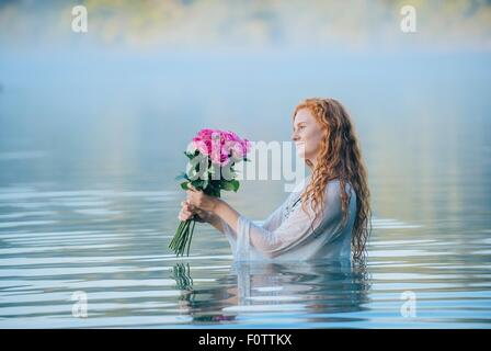 Young woman standing in lake gazing at bunch of pink roses Stock Photo