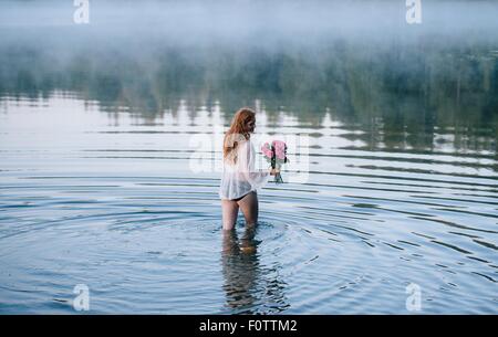 Rear view of young woman wading in misty lake holding bunch of pink roses Stock Photo