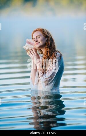 Young woman standing in misty lake with eyes closed listening to seashell Stock Photo