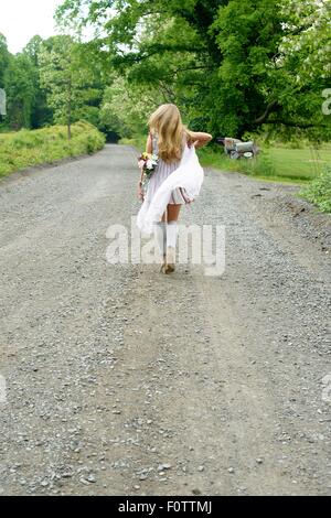 Rear view of young woman walking on rural road with flowers behind her back Stock Photo