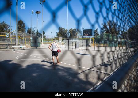 Young male basketball player behind court fence looking at basketball hoop Stock Photo