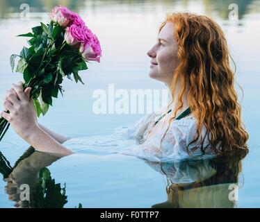 Head and shoulders of young woman with long red hair in lake gazing at bunch of pink roses Stock Photo