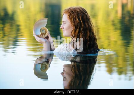 Head and shoulders of young woman with long red hair in lake gazing at seashell Stock Photo