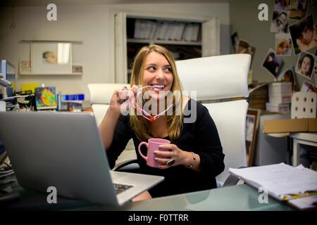 Young woman in office, sitting at desk, holding eyeglasses and coffee Stock Photo