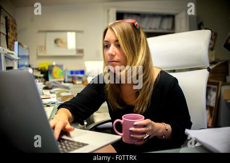 Young woman in office, sitting at desk, holding coffee cup, using laptop Stock Photo