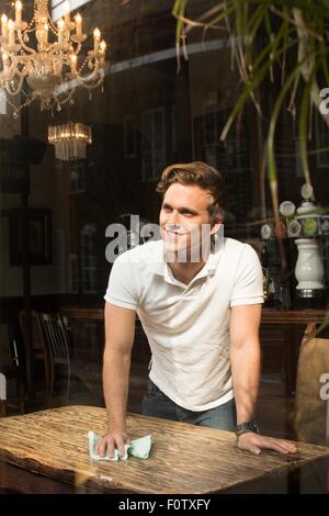 Young man cleaning table in public house, looking away smiling Stock Photo