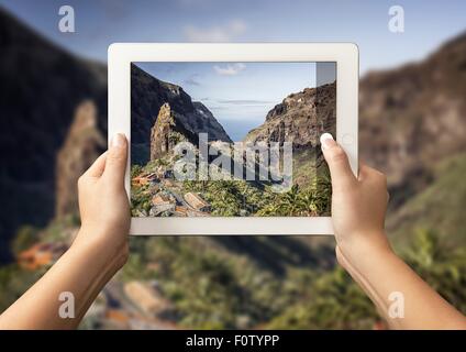 Hands of young woman holding up digital tablet with view of Masca Valley in front of view of Masca Valley, Tenerife, Spain Stock Photo