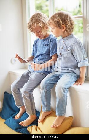 Two young brothers, sitting in window seat, looking at digital tablet Stock Photo