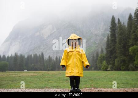 Portrait of female toddler wearing yellow raincoat in front of misty mountain, Yosemite National Park, California, USA Stock Photo